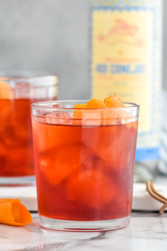 Photo of mezcal negroni cocktail in a tumbler with ice and garnished with an orange peel. A bottle of mezcal sits on the counter in the background.