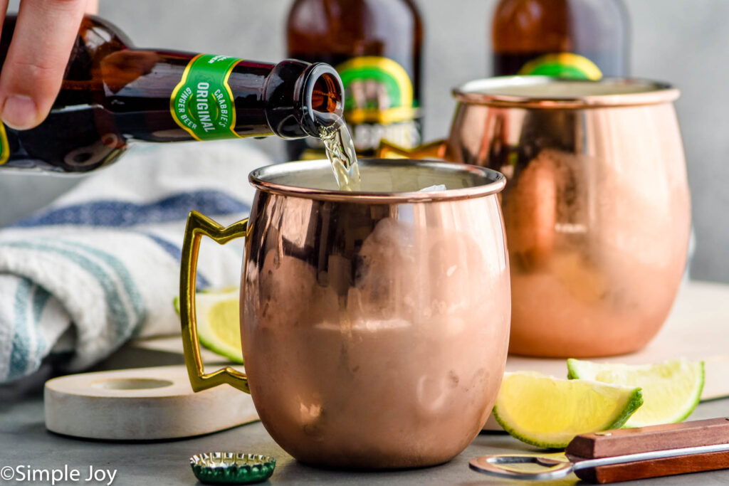 man's hand pouring bottle of ginger beer into a copper mug to make a moscow mule surrounded by bottles of ginger beer and lime wedges