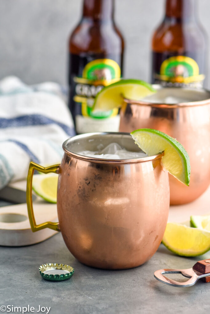 Two copper mugs of moscow mule recipe with ice and garnished with a lime wedge, two bottles of ginger beer sits in background