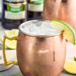 Two copper mugs of moscow mule recipe with ice and garnished with a lime wedge, two bottles of ginger beer sits in background