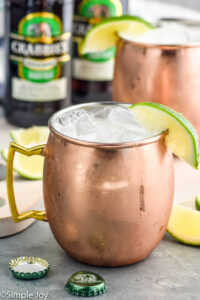 Two copper mugs of moscow mule recipe with ice and garnished with a lime wedge, two bottles of ginger beer sits in background