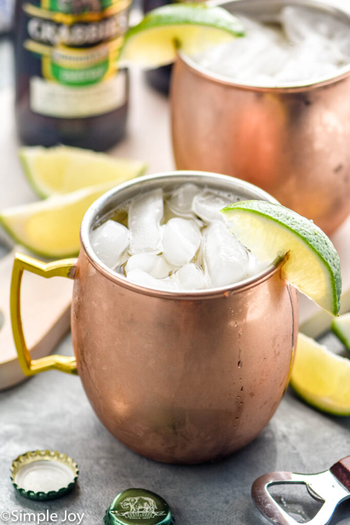 Two copper mugs of moscow mule recipe with ice and garnished with a lime wedge, bottle of ginger beer sits in background