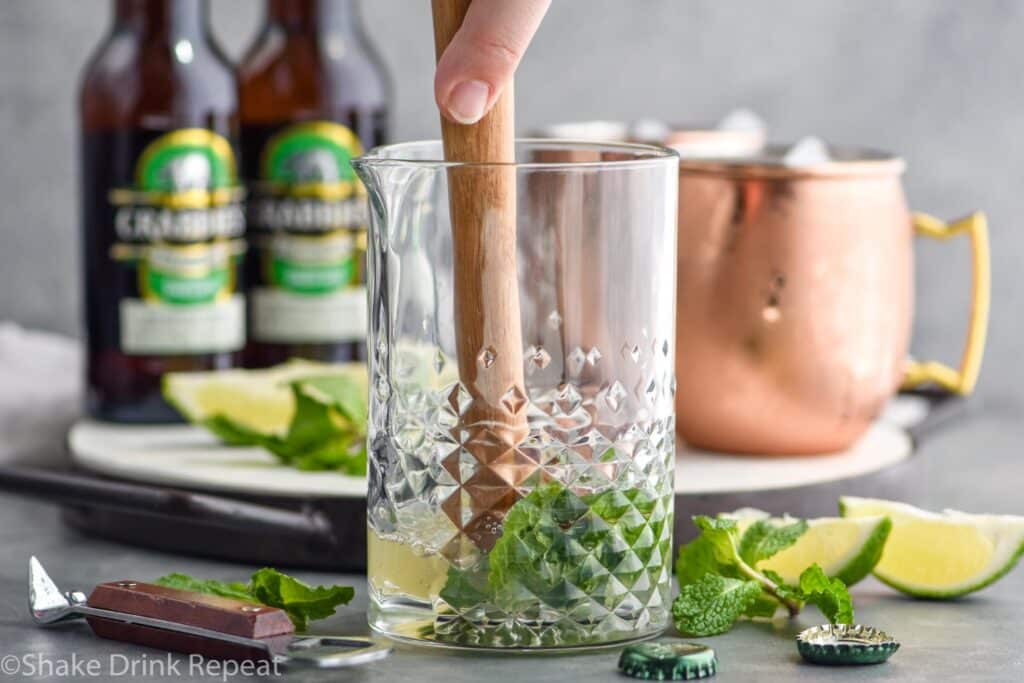man's hand muddling mint leaves with lime juice in a mixing glass to make a moscow mule with gin, with copper mugs, bottles of ginger beer, and lime wedges in the background