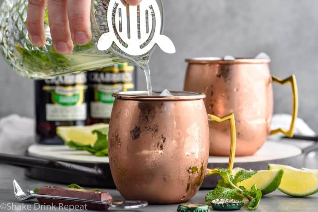 man's hand pouring mixing glass of muddled mint and lime juice into a copper mug of ice to make a moscow mule with gin, surrounded by fresh mint leaves, limes, and two bottles of ginger beer