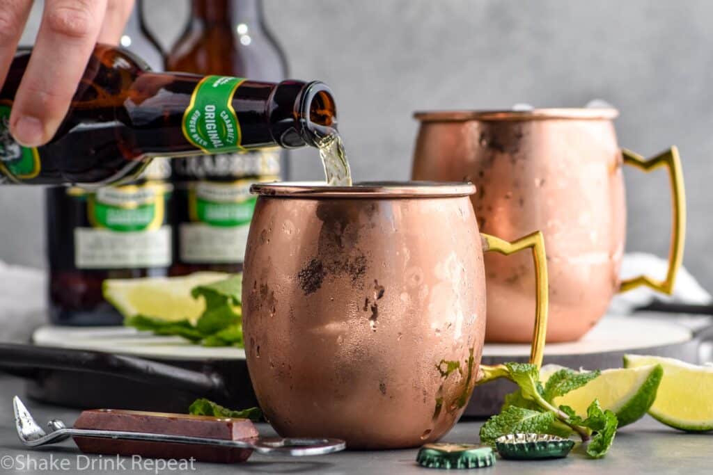 man's hand pouring bottle of ginger beer into a copper mug to make a moscow mule with gin, with bottles of ginger beer, lime wedges and fresh mint in background