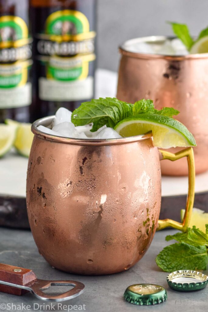 two copper mugs of moscow mule with gin with ice, garnished with mint leaves and limes, two bottles of ginger beer in background