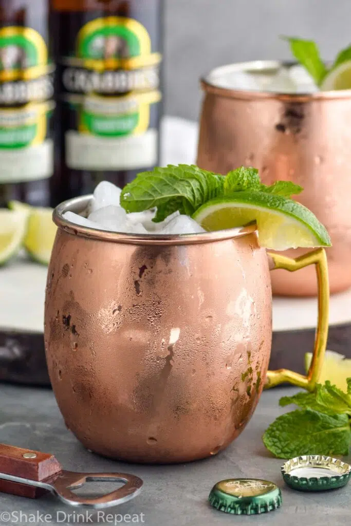 two copper mugs of moscow mule with gin with ice, garnished with mint leaves and limes, two bottles of ginger beer in background