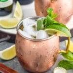 two copper mugs of moscow mule with gin with ice, garnished with mint leaves and limes, two bottles of ginger beer in background