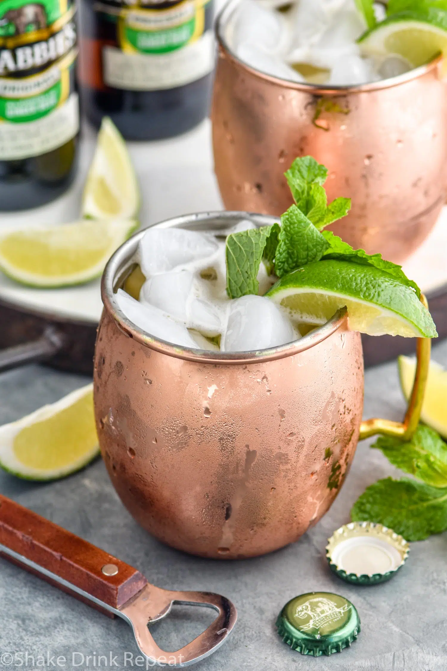 two copper mugs of moscow mule with gin with ice, garnished with mint leaves and limes, two bottles of ginger beer in background