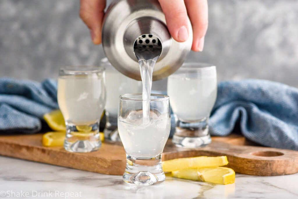 man's hand pouring cocktail shaker of white tea shot recipe into a shot glass with glasses of white tea shot and lemon slices in the background