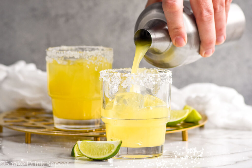 Photo of man's hand pouring Italian Margarita from a shaker bottle into a glass of ice with a salted rim. Lime wedges on the counter beside the glass.