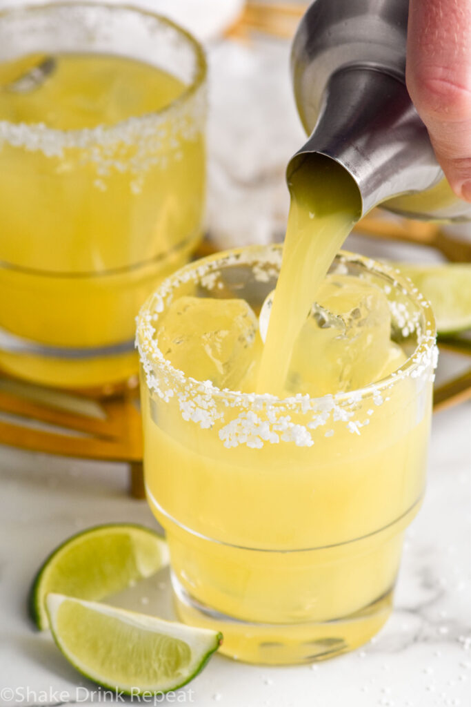 Overhead photo of man's hand pouring Italian Margarita from a shaker bottle into a glass of ice with a salted rim. Lime wedges on the counter beside the glass.