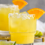 Photo of Italian Margarita recipe served in a glass with a salted rim and garnished with an orange slice. Lime wedges lay on the counter beside the glass.