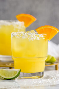 Photo of Italian Margarita recipe served in a glass with a salted rim and garnished with an orange slice. Lime wedges lay on the counter beside the glass.