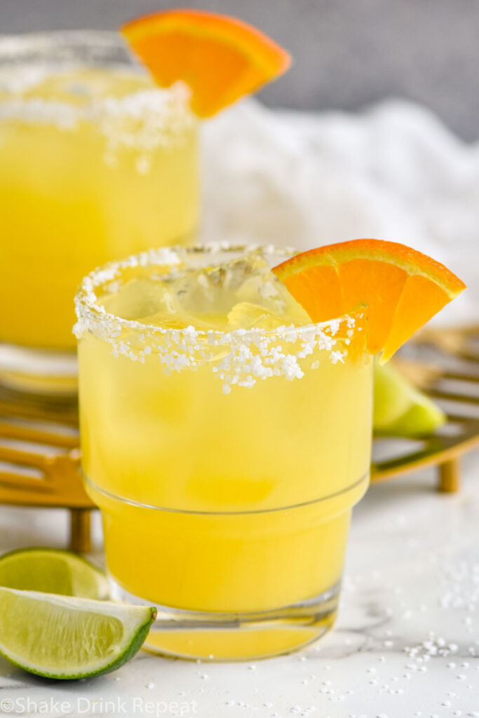 Overhead photo of Italian Margarita recipe served in a glass with a salted rim and garnished with an orange slice. Lime wedges lay on the counter beside the glass.
