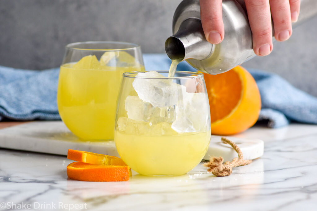 Photo of man's hand pouring Texas Margarita recipe from a shaker bottle into a glass of ice. A second Texas Margarita and a sliced orange sit on the counter in the background.
