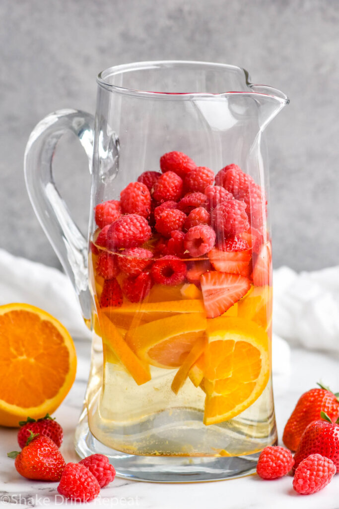 glass pitcher of white sangria ingredients surrounded by fresh orange, strawberries and raspberries