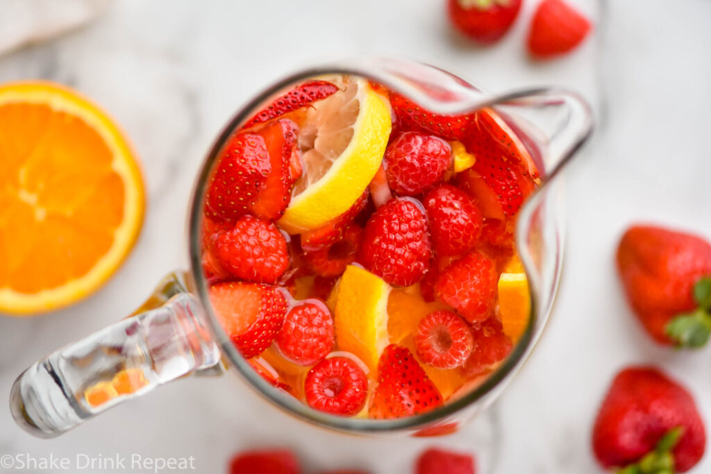 overhead view of glass pitcher of white sangria surrounded by fresh orange, strawberries and raspberries