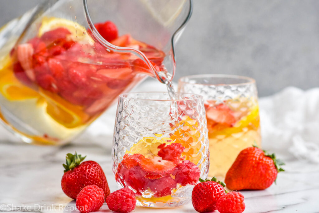 glass pitcher of white sangria pouring into a glass surrounded by fresh strawberries and raspberries