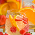 glass of white sangria recipe garnished with an orange slice, fresh strawberry and raspberries laying beside glass
