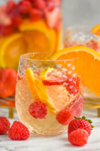 glass of white sangria recipe garnished with an orange slice, fresh strawberry and raspberries laying beside glass