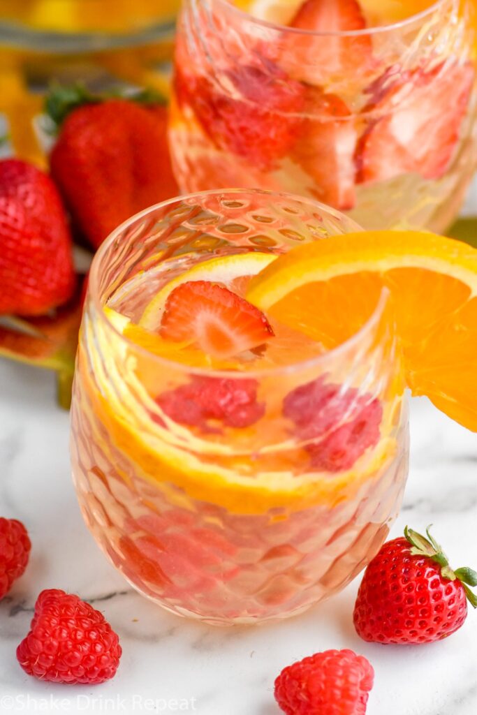 glass of white sangria recipe garnished with an orange slice, fresh strawberry and raspberries laying beside glass