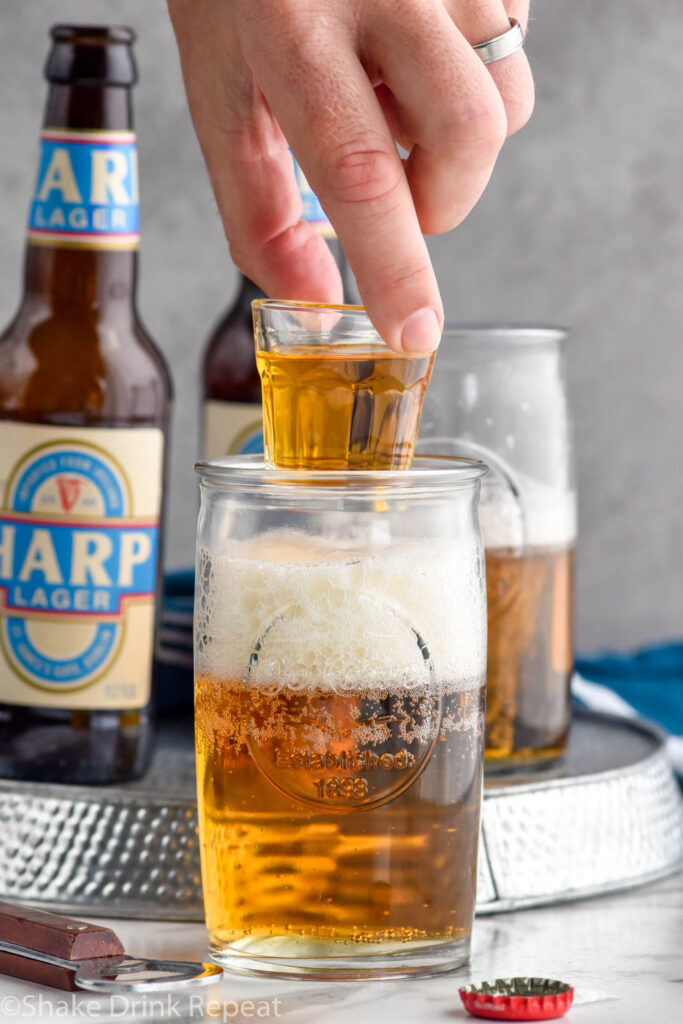 Man's hand holding shot glass of whiskey over a glass of beer to make a Boilermaker drink. Two bottles of lager beer and glass sit in background