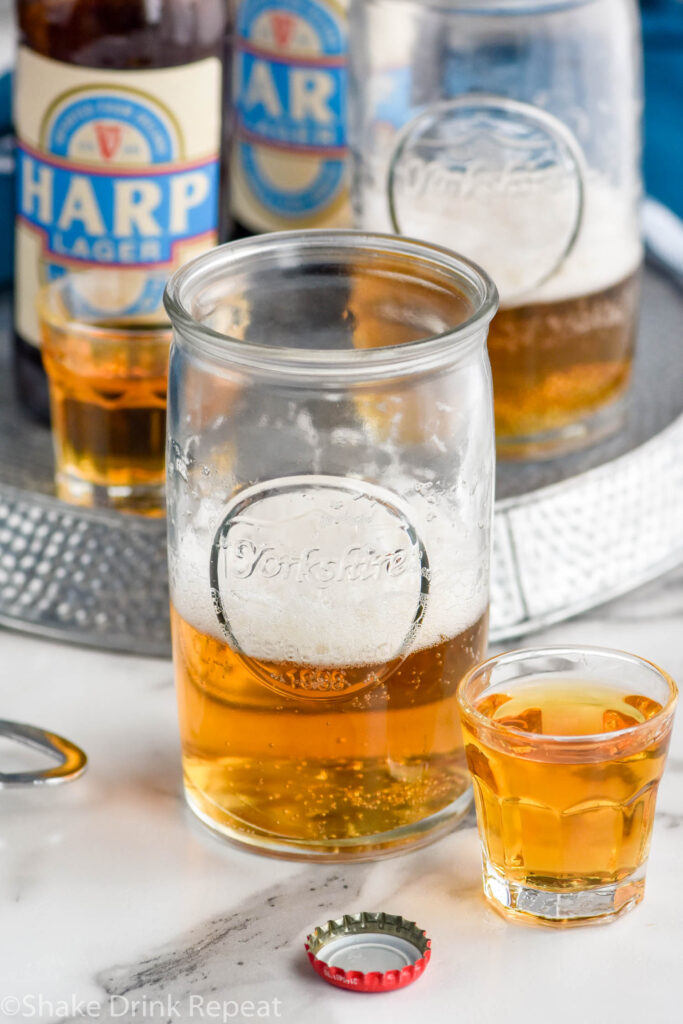 Glass of beer next to shot of whiskey to make a Boilermaker drink with bottles of beer and shot glass of whiskey