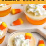 Pinterest graphic of candy corn jello shots. Text says "vodka candy corn jello shots shakedrinkrepeat.com" Image shows multiple shot glasses of candy corn jello shots topped with whipped cream and surrounded by candy corn candy