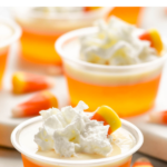Pinterest graphic of candy corn jello shots. Text says "the best candy corn jello shots shakedrinkrepeat.com" Image shows multiple shot glasses of candy corn jello shots topped with whipped cream and surrounded by candy corn candy