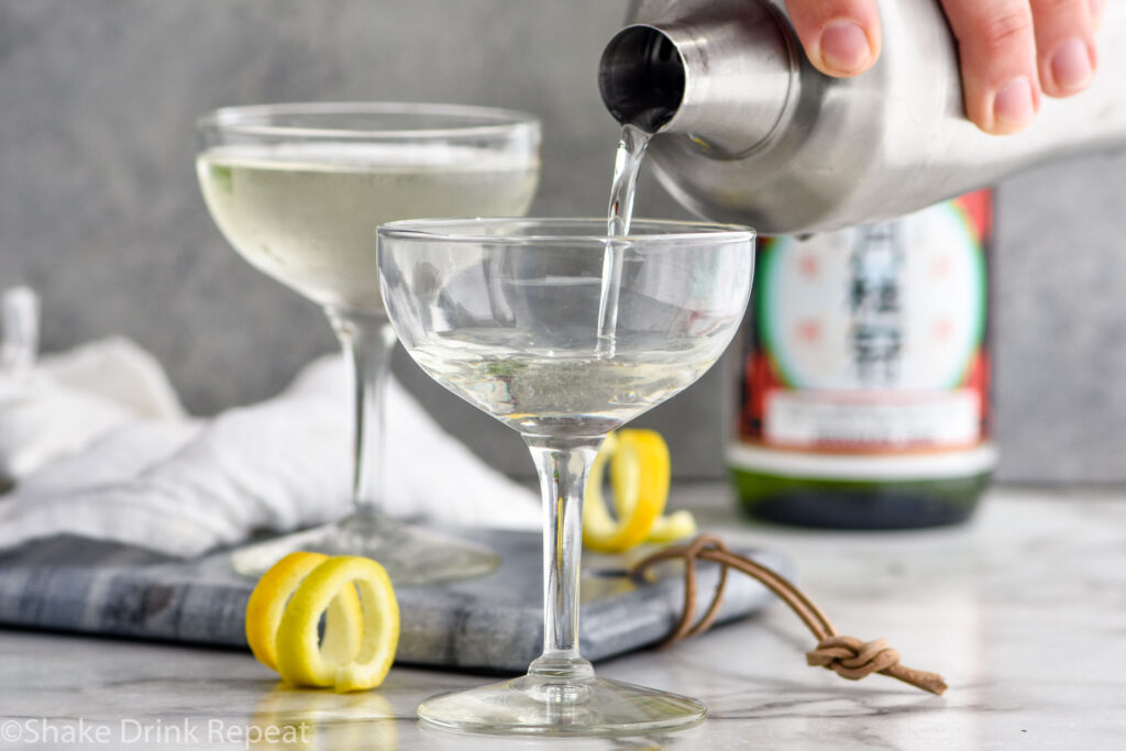 man's hand pouring cocktail shaker of Saketini ingredients into a glass with glass of Saketini, bottle of sake, and lemon twist in background