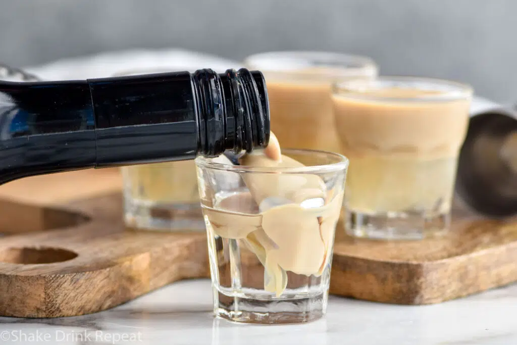 bottle of Bailey's Irish cream pouring over the back of a spoon into a shot glass of Sambuca to make a slippery nipple shot recipe, shot glasses of slippery nipple sit in background