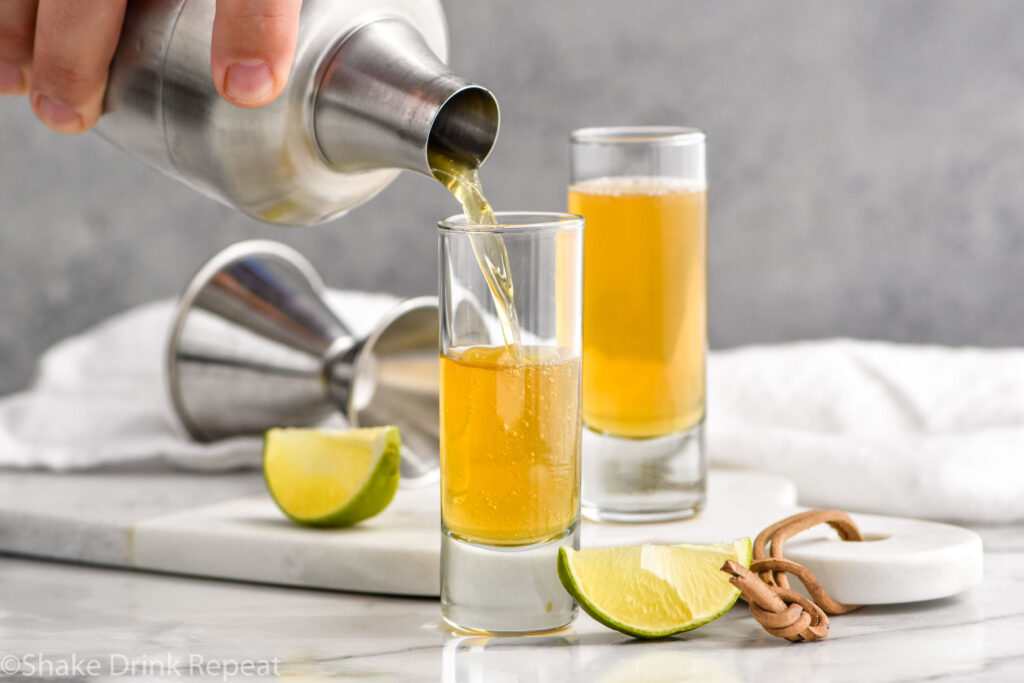 man's hand pouring cocktail shaker of snakebite shot ingredients into a shot glass. Lime wedges and shot glass of snakebite shot sit in background