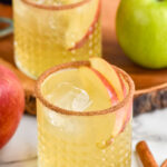 two glasses of apple cider margaritas with ice, apple slices, and cinnamon sugar rim. Cinnamon sticks, red apple and green apple surrounding.