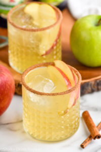 two glasses of apple cider margaritas with ice, apple slices, and cinnamon sugar rim. Cinnamon sticks, red apple and green apple surrounding.