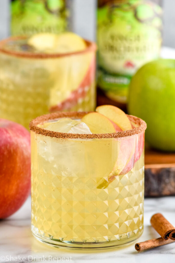 glass of apple cider margarita with ice, apple slices, and cinnamon sugar rim with two bottles of angry orchard hard cider, green and red apples, and glass of apple cider margarita in background.