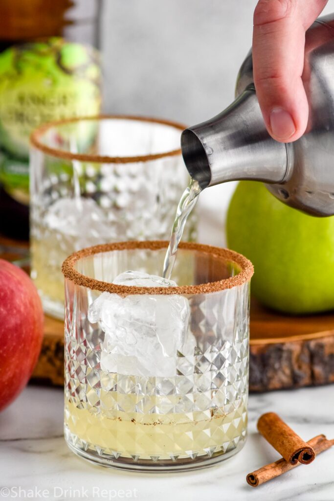 man's hand pouring cocktail shaker of apple cider margaritas ingredients into a glass of ice with cinnamon sugar rim. Bottle of angry orchard hard cider, green apple, red apple, and cinnamon sticks in background.