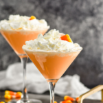 Pinterest graphic of candy corn martini recipe. Text says "the best candy corn martini only uses 2 ingredients! shakedrinkrepeat.com" Image shows two martini glasses of candy corn martini topped with whipped cream and candy corn pieces. Candy corn pieces surround base of glasses