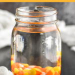 Pinterest graphic of Candy Corn Vodka. Text says "candy corn vodka so easy! shakedrinkrepeat.com" Image shows a mason jar of vodka and candy corn to make candy corn vodka. Candy corns surrounding jar.
