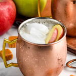 copper mug of caramel apple moscow mule with ice and garnished with apple slices. Fresh apples and caramels sit in background and surrounding.