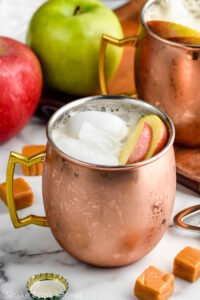 copper mug of caramel apple moscow mule with ice and garnished with apple slices. Fresh apples and caramels sit in background and surrounding.