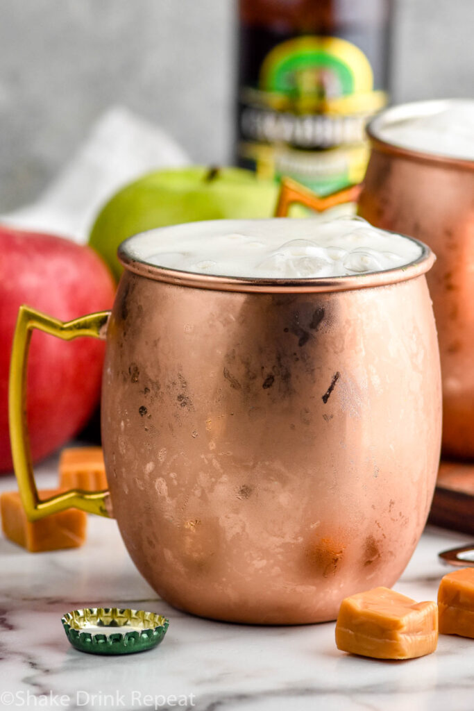 copper mug of caramel apple moscow mule with ice and garnished with apple slices. Fresh apples, caramels, and bottle of ginger beer sit in background and surrounding.