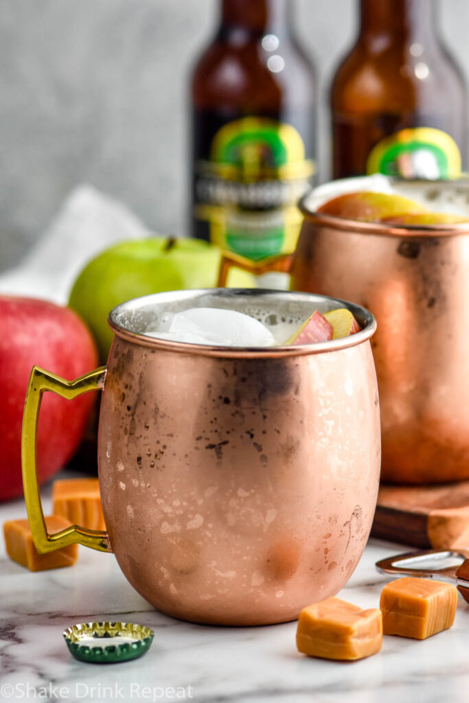two copper mugs of caramel apple moscow mules with ice and garnished with apple slices. Bottle of ginger beer, apples, and caramels sit in background