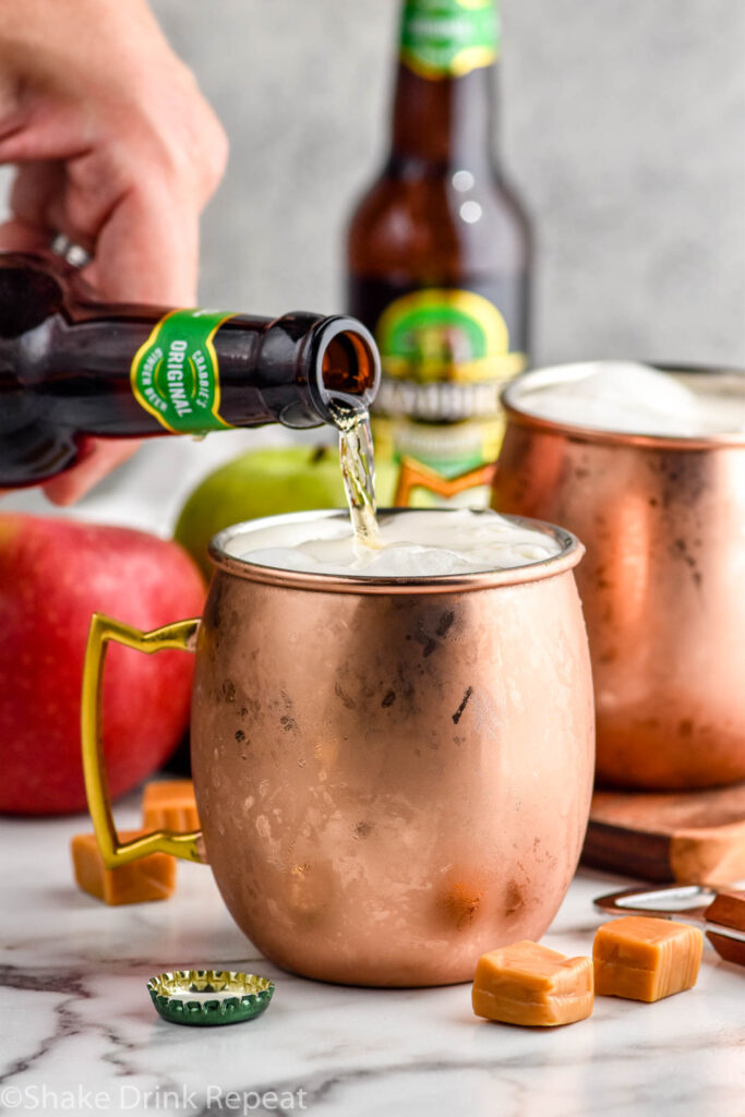 man's hand pouring bottle of ginger beer into a copper mug of caramel apple moscow mule ingredients. Mug of caramel apple moscow mule, apples, and caramels sit beside and behind.