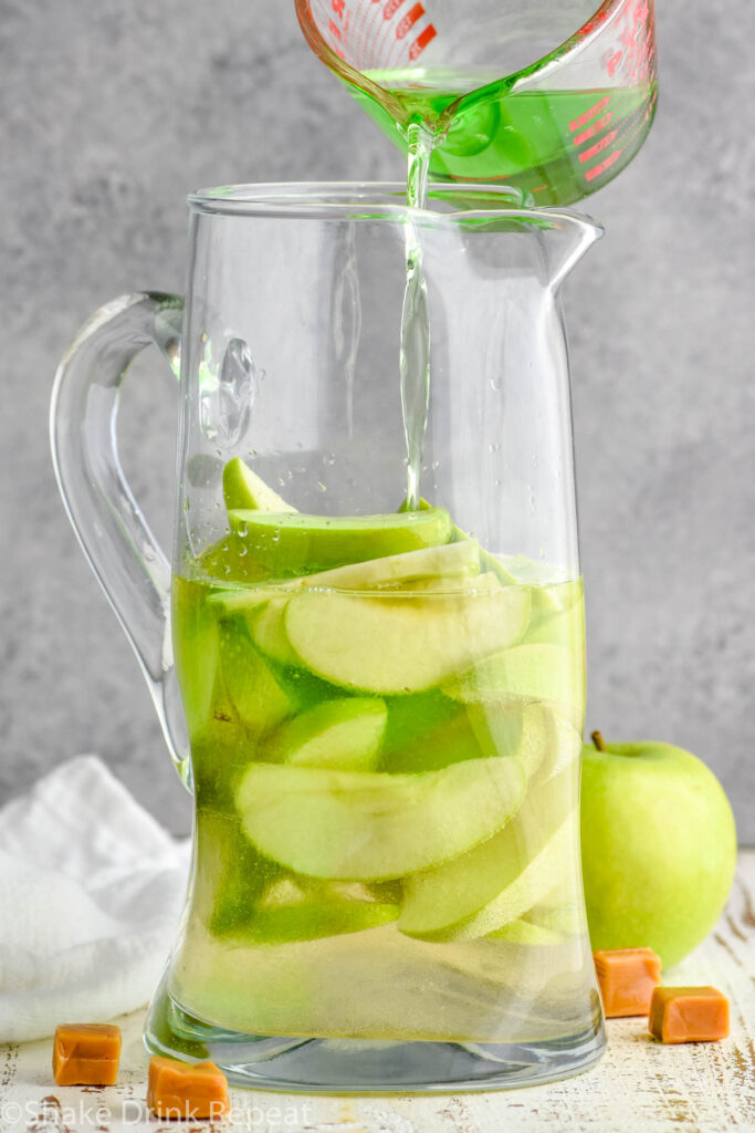 measuring glass of apple liqueur pouring into a glass pitcher of caramel apple sangria with slices of fresh apples surrounded by a green apple and caramel candies