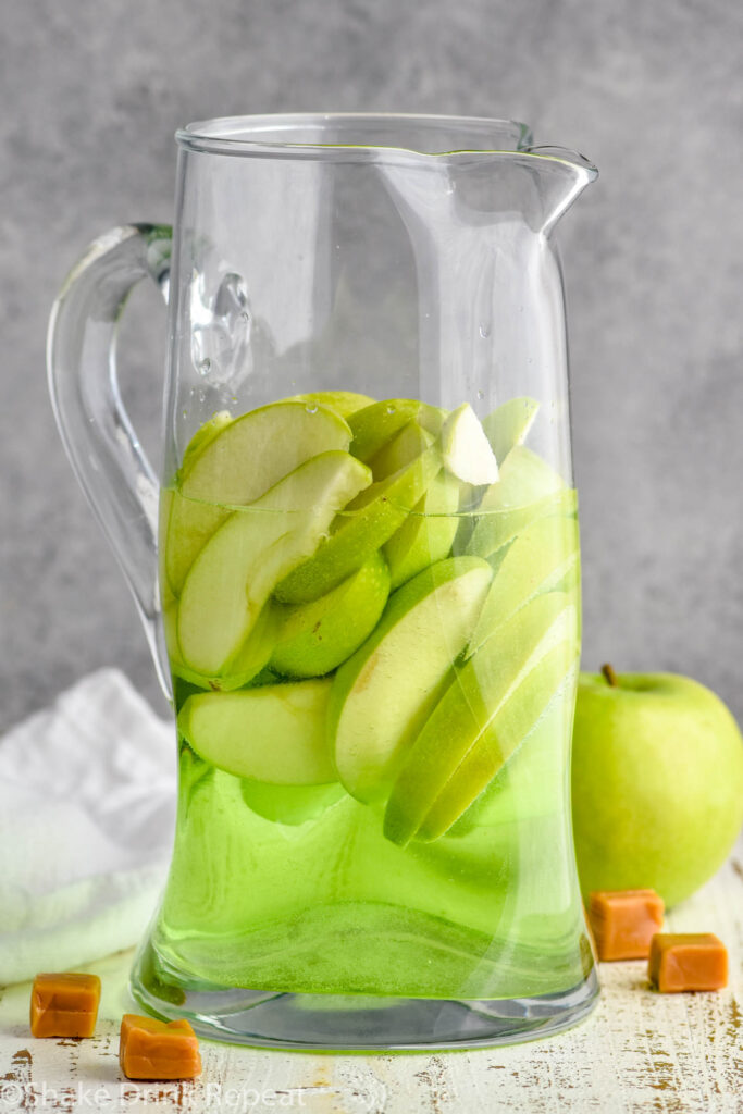 glass pitcher of caramel apple sangria recipe with fresh apples and caramel candies surrounding.