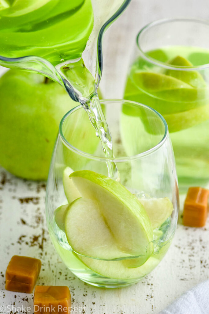 pitcher of caramel apple sangria pouring into a glass of fresh apple slices surrounded by caramel candies