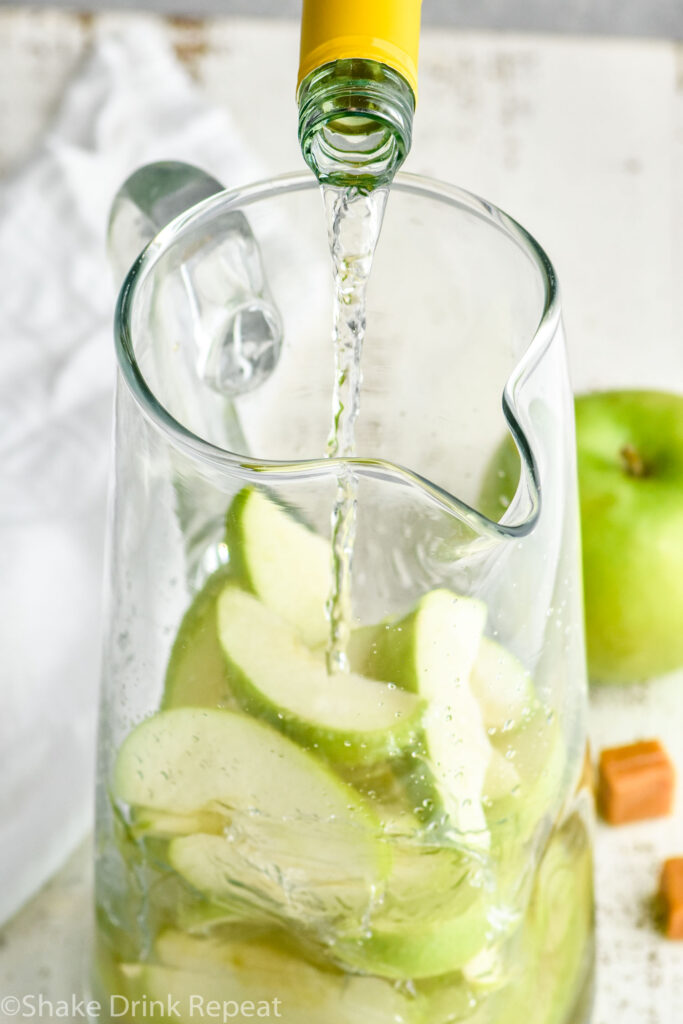 bottle of white wine pouring into a glass pitcher of apple slices and caramel apple sangria ingredients with green apple and caramel candies in background