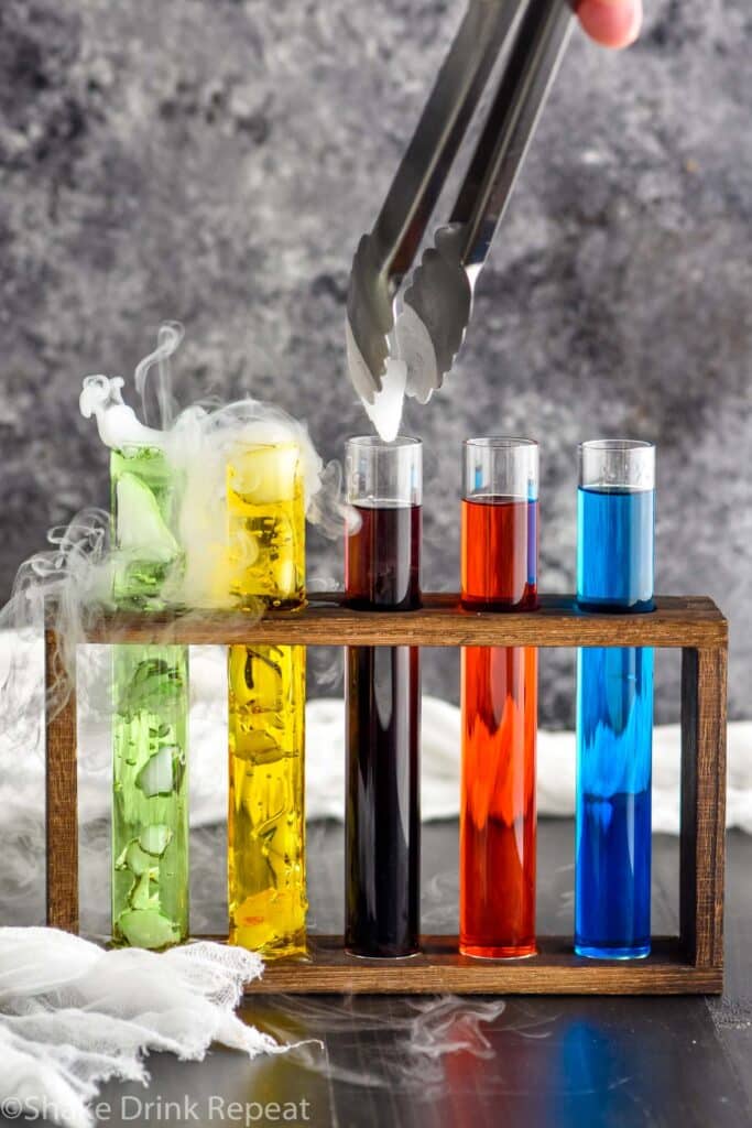tongs dropping piece of dry ice into a test tube of mad scientist shot surrounded by other test tubes of mad scientist shots in a wooden holder