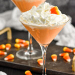 Two martini glasses of candy corn martini topped with whipped cream and garnished with candy corn candy. Candy corn surrounding base of glasses.